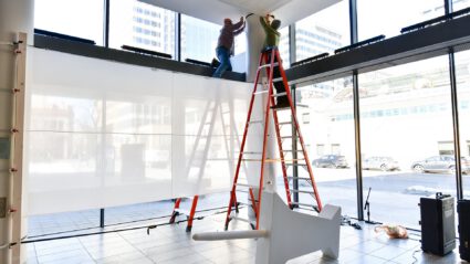 Two workers install large panels on a wall inside a modern building with floor-to-ceiling windows.