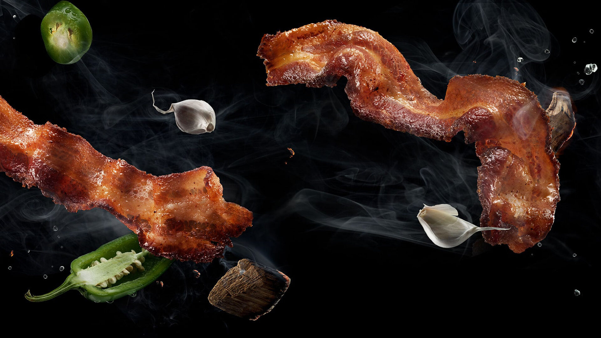 Crispy bacon strips, garlic, and jalapeño peppers floating in smoke against black background.