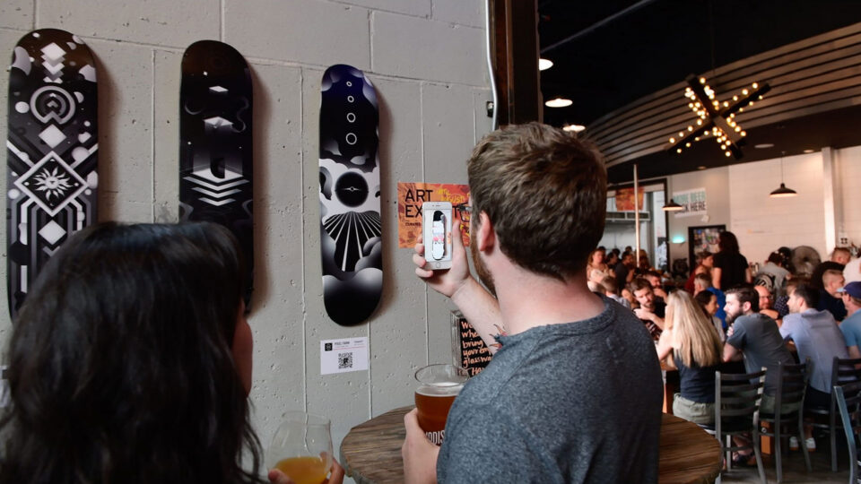 Man at bar scanning QR code on wall with phone, surrounded by patrons and skateboards.