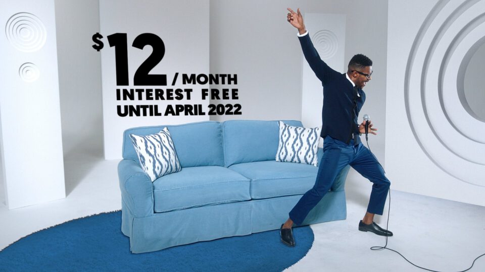 Man joyfully sings into mic beside blue sofa with $12/month interest-free offer