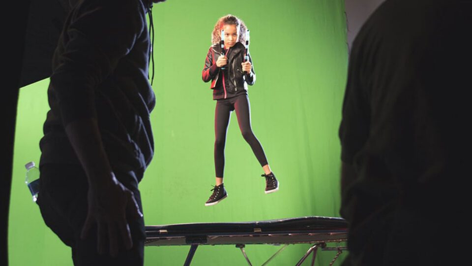 Young girl in black outfit poses on green screen set with crew around