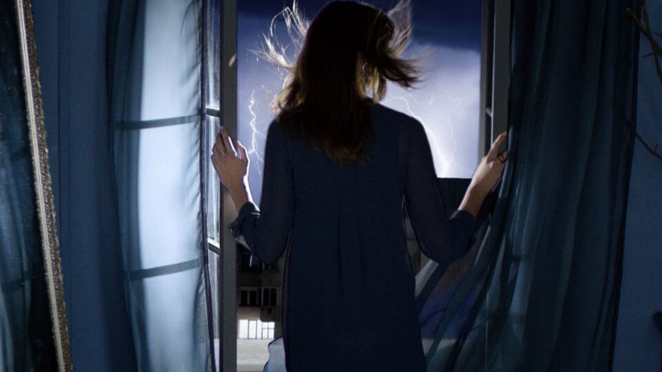 Woman stands at window, curtains parted, lightning flashes outside in stormy night.
