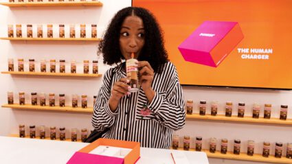 Woman sips from a can in a store with shelves of products and a screen reading "The Human Charger"