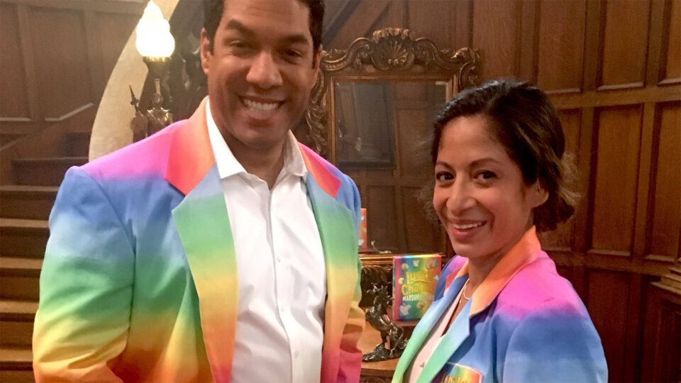 Two people in colorful rainbow jackets smile in a wood-paneled room.