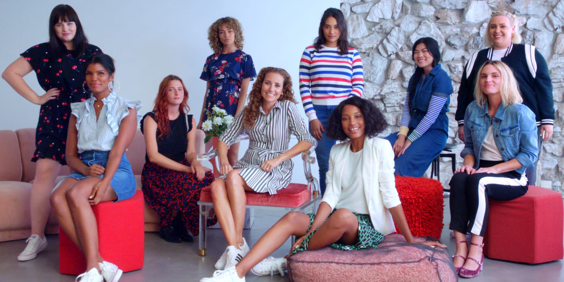 Nine diverse women in stylish outfits posing together in a modern room with red stools and stone wall.