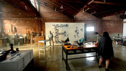Artists work in a spacious brick studio with exposed beams and creative clutter.