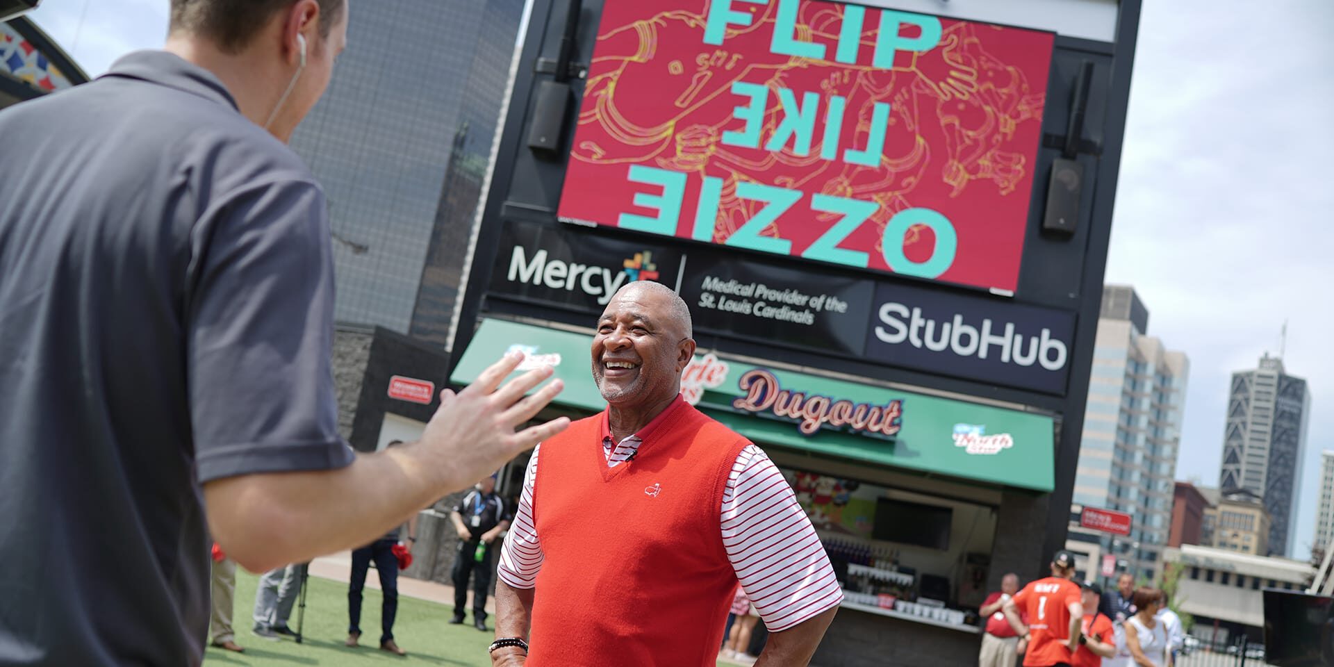Man in red vest smiles while interacting with another person near stadium scoreboard.