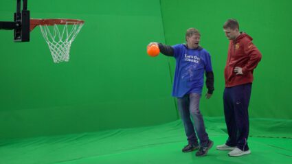 Two men in front of green screen, one holding orange ball near basketball hoop.