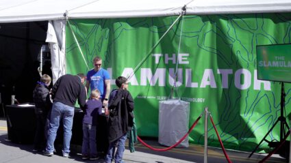 People gather around a green tent promoting "The Slamulator" tech product.