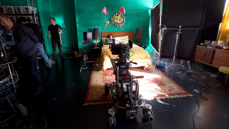Film crew shoots scene in vibrant green bedroom with camera on dolly.