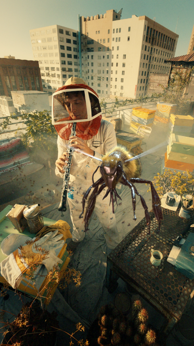 Person in beekeeper gear plays flute with giant bee hovering nearby on rooftop