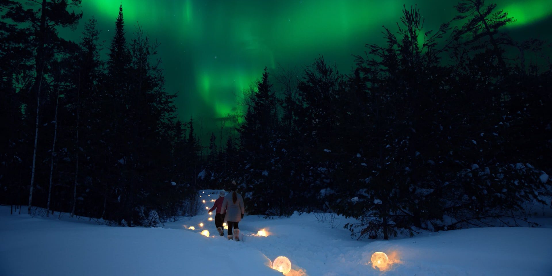 Two people walk through snowy forest under green aurora borealis with glowing orbs.