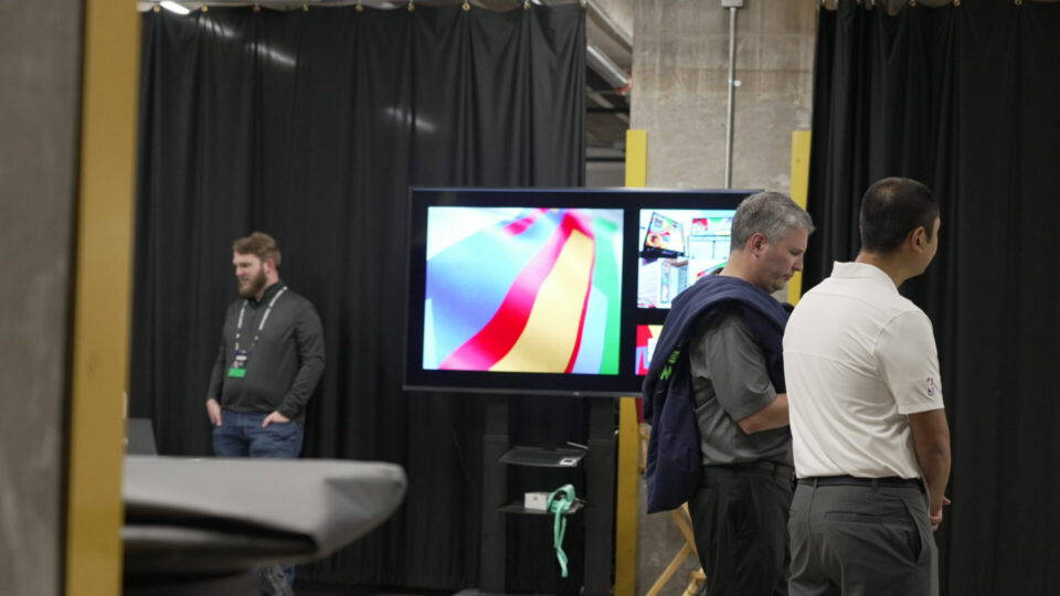 Three men observe a large display screen at an indoor event.
