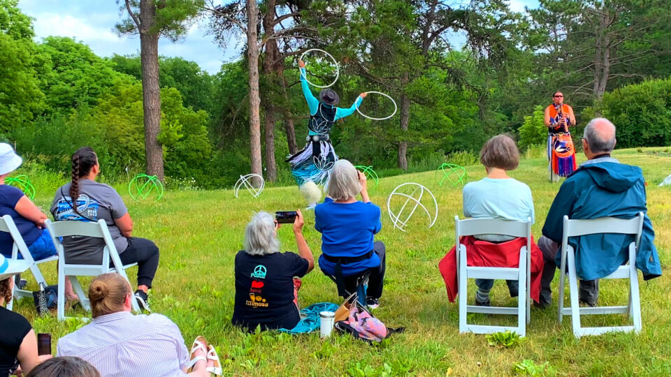 Person performs hoop dance outdoors as audience watches from chairs