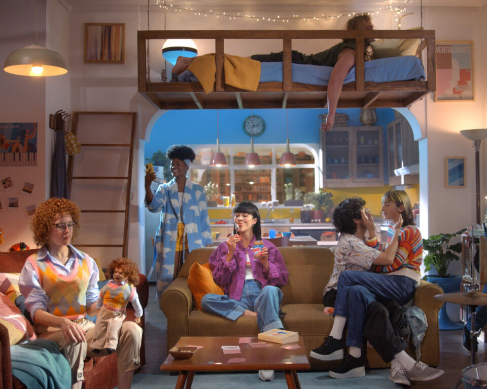 Six diverse people socializing in a colorful, loft-style living space with string lights and a mezzanine bed.