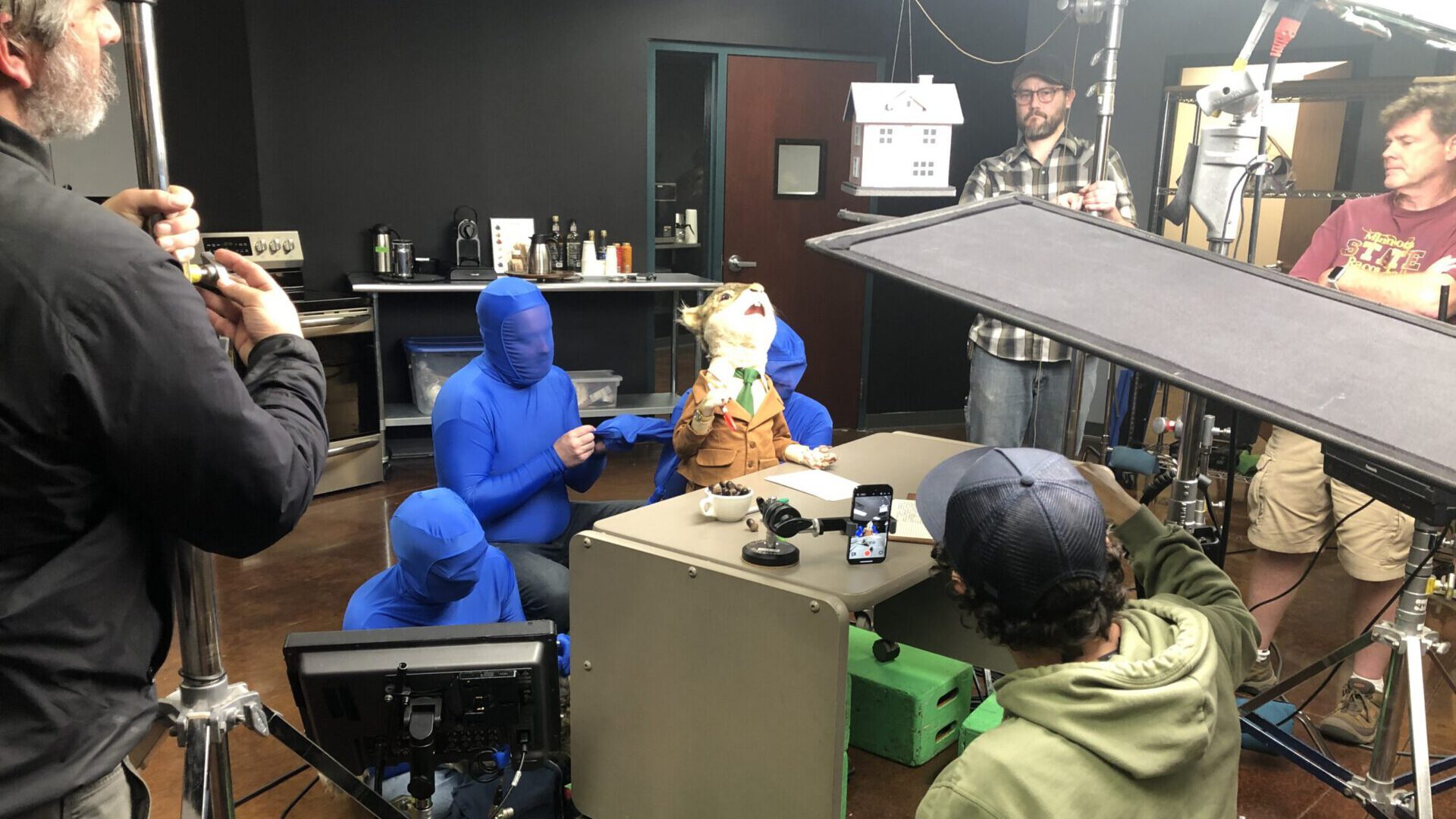 Behind-the-scenes shot of a film set with crew, blue-suited actors, and a puppet at a desk.