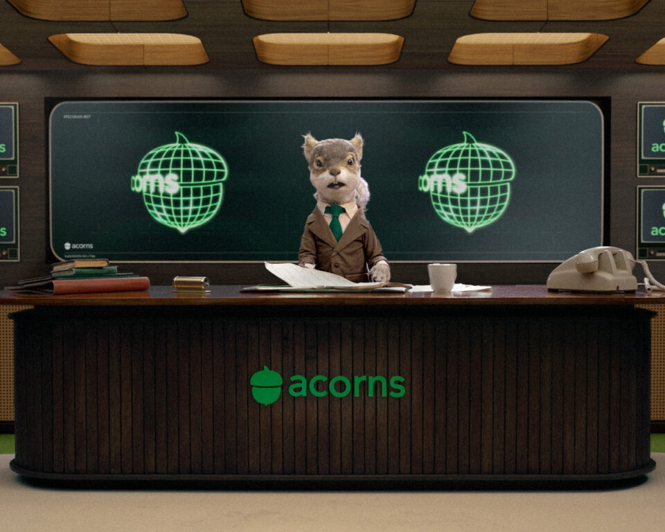 Squirrel puppet in suit at Acorns desk with glowing acorn logos