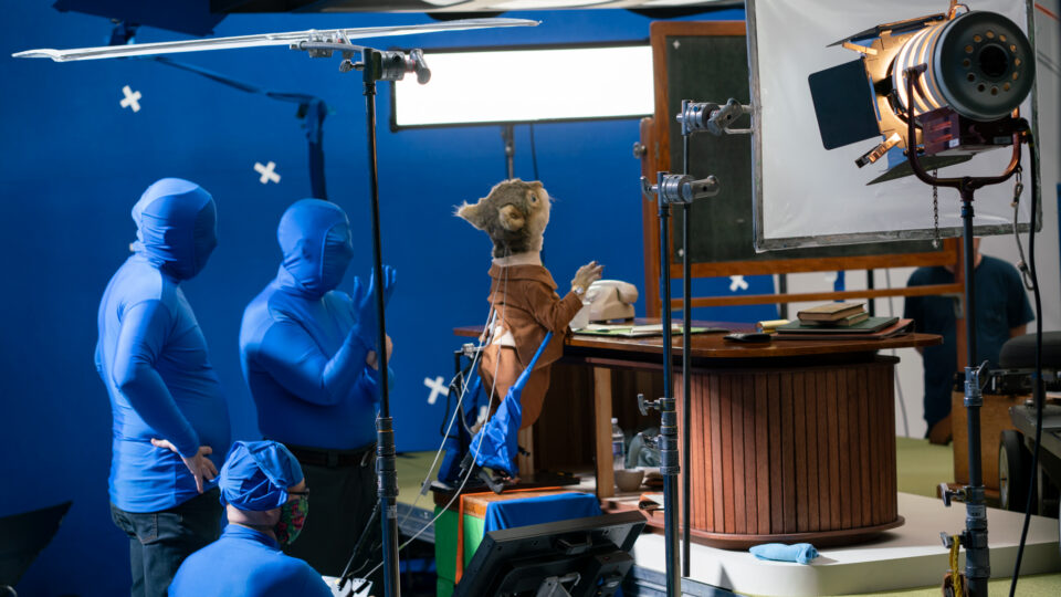 Puppet studio scene with blue-suited crew and a wooden desk setup.