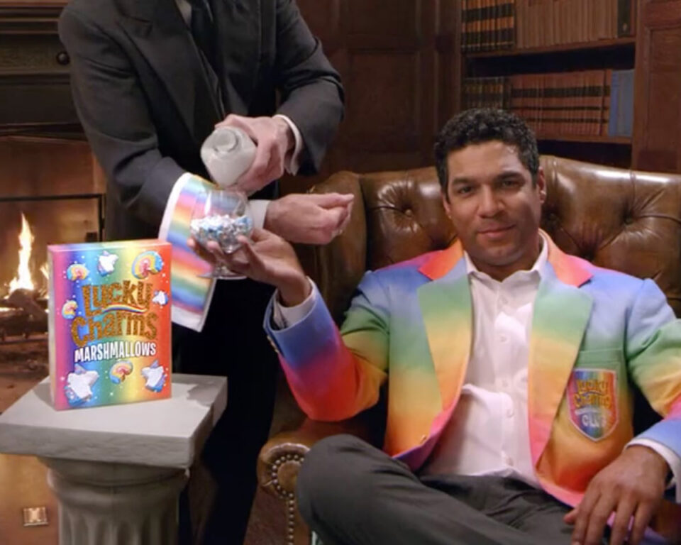 Man in rainbow suit receives marshmallows from server beside Lucky Charms box.