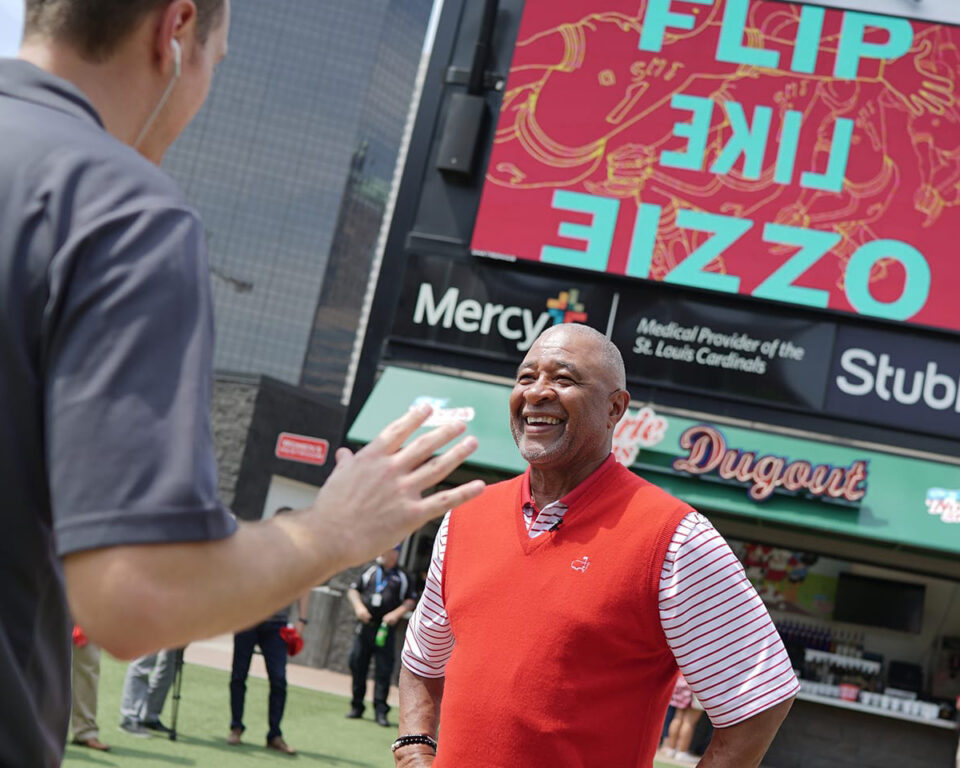 Ozzie Smith in red vest waves at another man near stadium signage