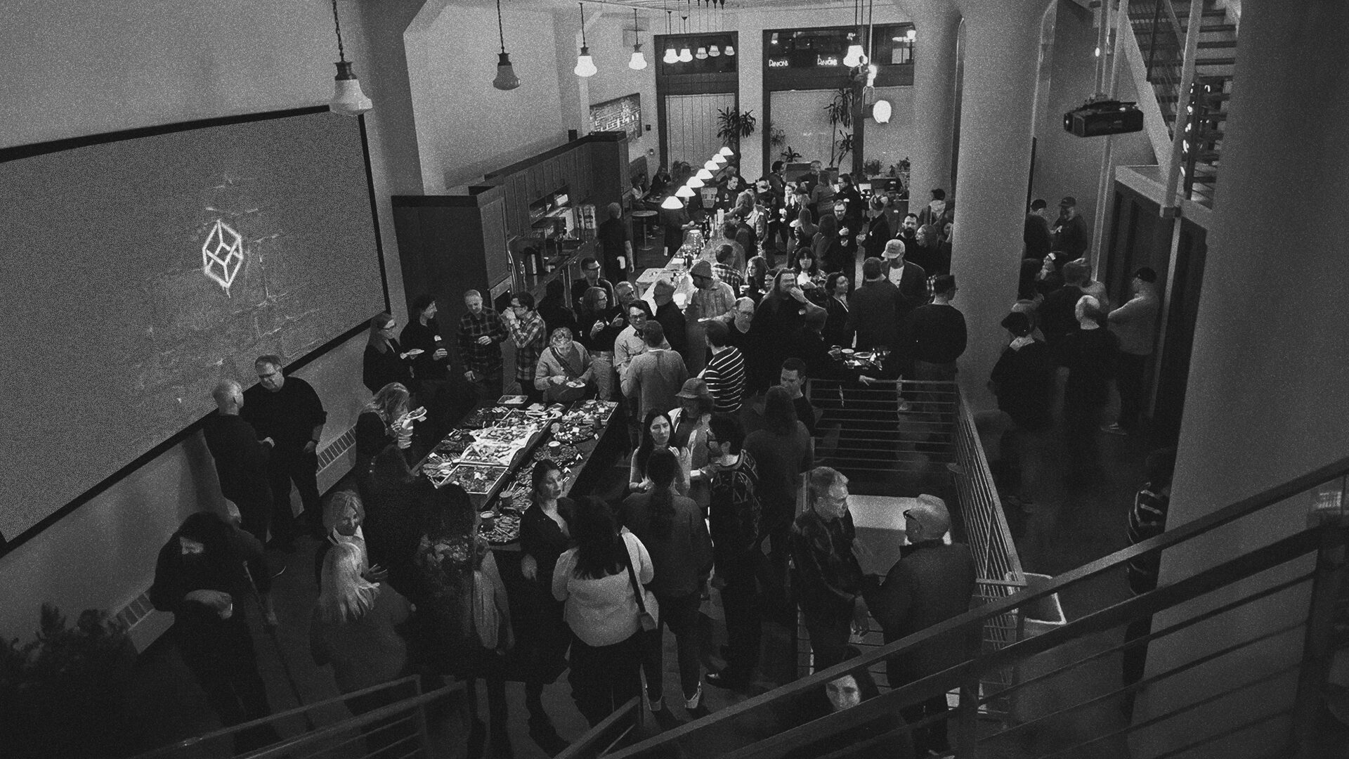 Black-and-white photo of a crowded indoor event with people mingling near food tables and stairs.