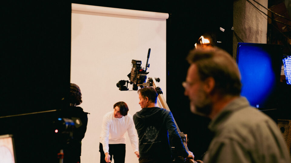 Film crew shoots scene with actor in white shirt against white backdrop