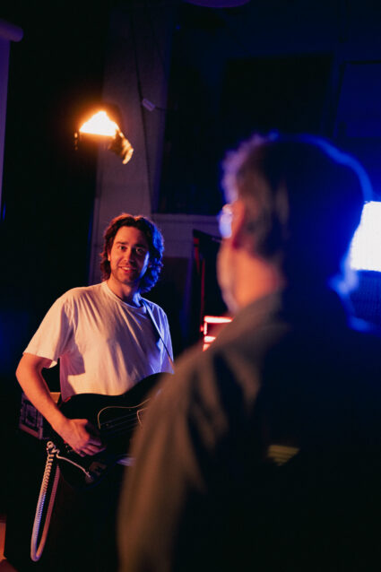Musician with guitar smiles at viewer in dim, colorful studio lighting.