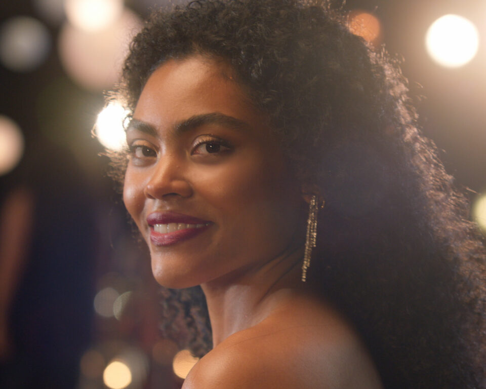Woman with curly hair smiles warmly against blurred lights