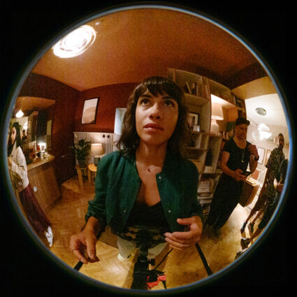 Fisheye lens captures woman in green jacket looking up in warmly lit indoor space with people around.