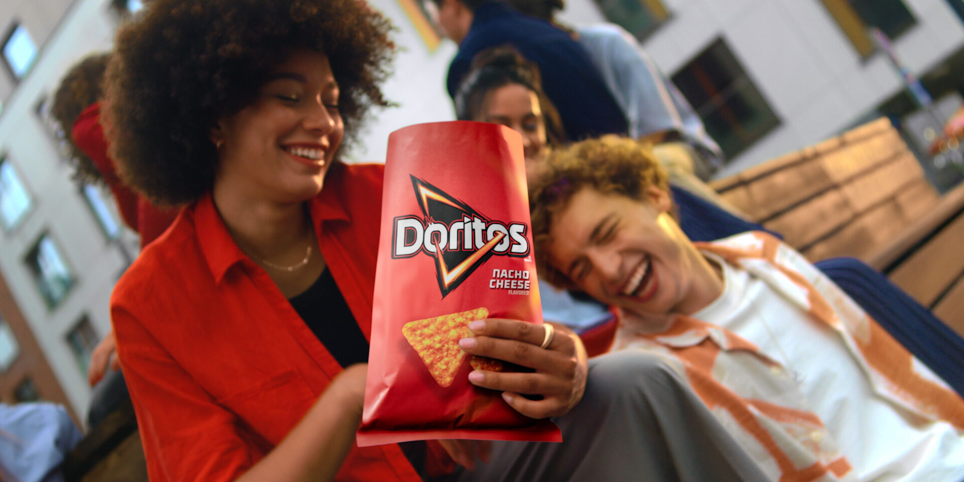 Two smiling friends share a bag of Doritos Nacho Cheese chips outdoors.