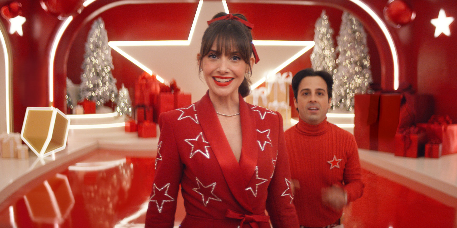 Alison Brie smiles in a red star-studded suit on a stylized red set for a Macy's holiday campaign.