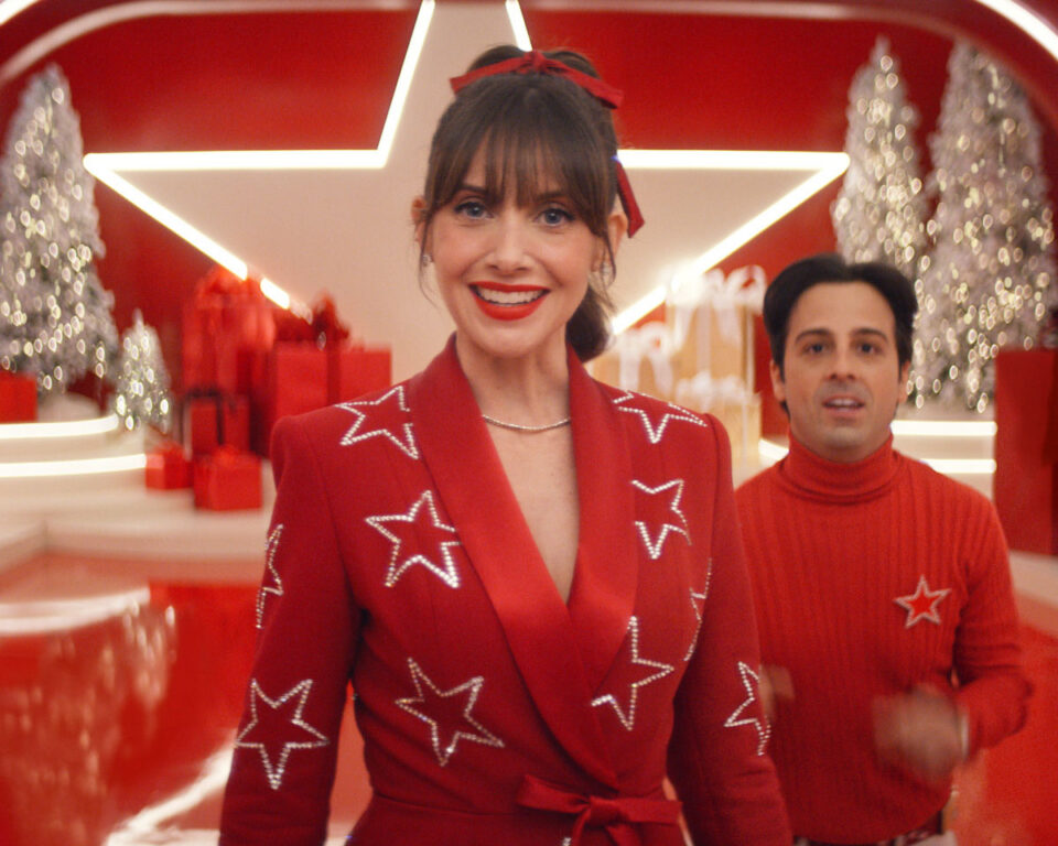 Alison Brie smiles in a red star-studded suit on a stylized red set for a Macy's holiday campaign.