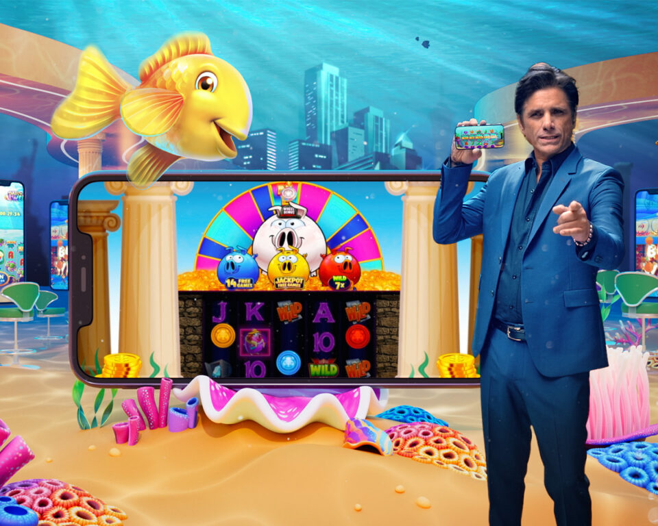 John Stamos in blue suit holds phone showing colorful underwater slot game with fish and symbols.