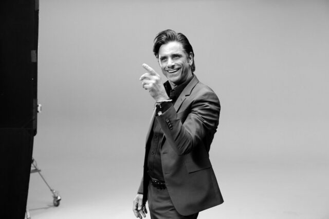 John Stamos in suit pointing and smiling in studio setting