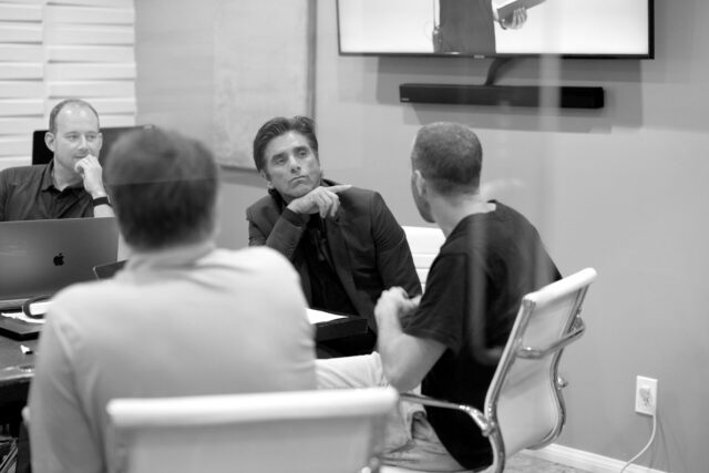 John Stamos and three people in a meeting room, one with laptop, others engaged in discussion