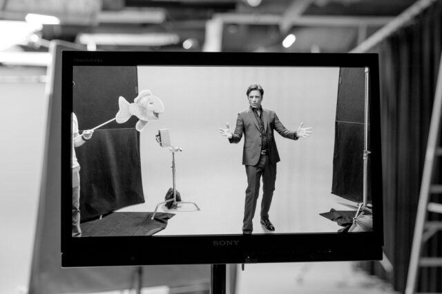 John Stamos in suit gestures on film set with puppet fish visible