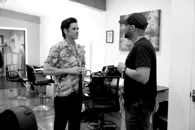 John Stamos and man converse in a studio; one adjusts his shirt, the other listens attentively.