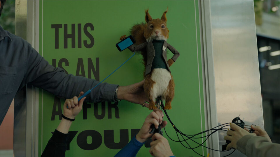 Squirre puppet l in suit held by humans with wires and phone against green sign