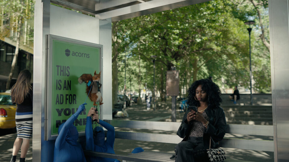Person in blue suit holds squirrel puppet near bus stop ad for Acorns.