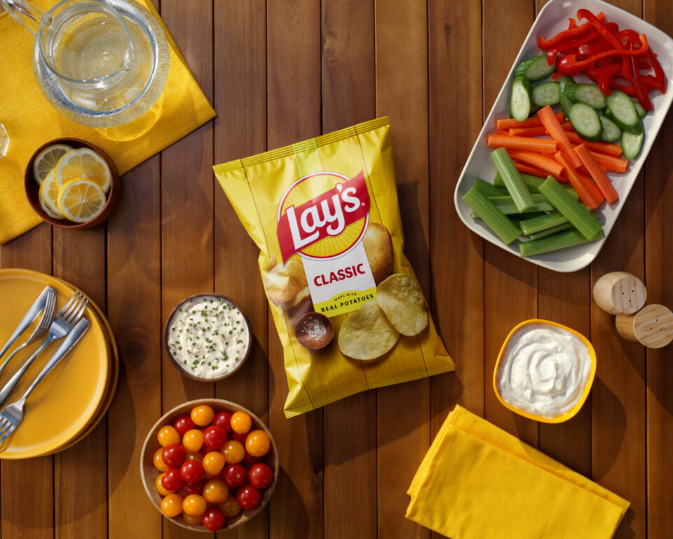Lay's Classic potato chips bag surrounded by fresh veggies, creamy dipping sauce, and bright yellow tableware on a wooden table.