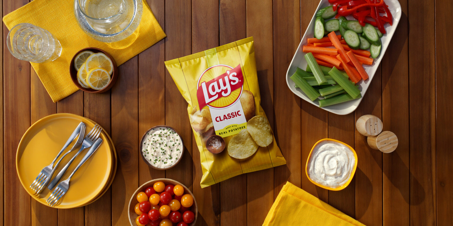 Lay's Classic potato chips bag surrounded by fresh veggies, creamy dipping sauce, and bright yellow tableware on a wooden table.